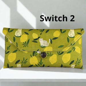 May include: A rectangular, lime green clutch bag with a flap closure. The fabric features a pattern of yellow lemons, green leaves, and white cats. The bag is secured with a brown button. The text "Switch 2" is printed on the top.