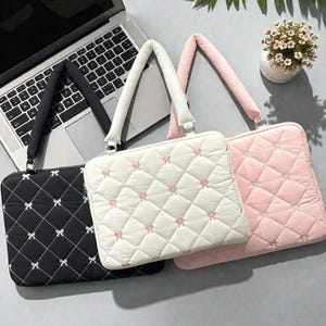 May include: Three quilted laptop cases in black, white, and pink. The black case has a white bow pattern, the white case has pink bows, and the pink case has a quilted pattern. Each case has a matching handle.