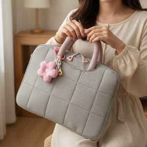 May include: A gray quilted laptop bag with a pink flower charm. The bag has a soft, padded appearance with a square pattern. The handles are a light pink color, and the zipper pulls are pink. The bag is designed to carry a laptop or other electronic devices.