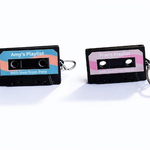 Könnte beinhalten: Zwei schwarze Kassettenband-Schlüsselanhänger. Einer hat ein blau-oranges Etikett mit "Amy's Playlist" und "with love from Pete". Der andere hat ein rosa Etikett mit "Fran's Playlist" und "with love from Marie".