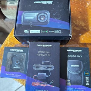 Op de afbeelding: Een verzameling Nextbase Dash Cam-producten, waaronder een hoofdeenheid, een achteruitrijcamera, een hardwire kit en een 32GB Go Pack. De dozen zijn zwart met witte en blauwe tekst en bevatten productafbeeldingen en branding.