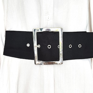 May include: A wide black belt with a large, square silver buckle. The belt is worn over a white button-down shirt. The buckle has a rectangular shape and is the focal point of the belt's design.