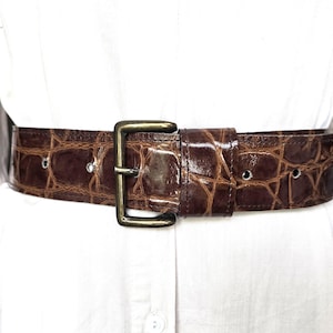 Op de afbeelding: Een brede bruine riem met een glanzende krokodil-reliëftextuur en een rechthoekige metalen gesp in antieke messingkleur. De riem wordt gedragen over een effen wit overhemd en heeft zichtbare metalen oogjes langs de band voor verstelling.