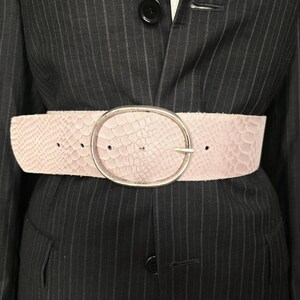 Puede incluir: Un cinturón ancho de color rosa claro con un patrón texturizado de piel de serpiente y una gran hebilla ovalada de metal plateado. El cinturón se lleva sobre una americana gris oscuro a rayas con botones negros visibles.