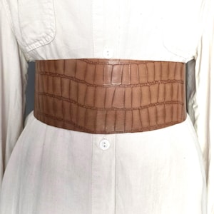 Puede incluir: Cinturón ancho marrón de piel sintética con textura de piel de cocodrilo. El cinturón se lleva sobre una camisa blanca abotonada. El cinturón mide aproximadamente 15 cm de ancho.