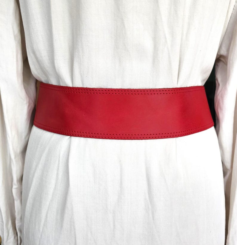 May include: A wide, bright red leather belt worn over a white garment. The belt is a solid color with a stitched border. The belt is a wide style, designed to cinch the waist. The garment is a light, flowing fabric.