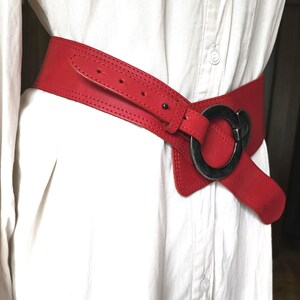 May include: A wide, red leather belt with a large, circular, silver-toned buckle. The belt is worn over a white shirt, showcasing its bold design and color. The belt's width is approximately 4 inches.