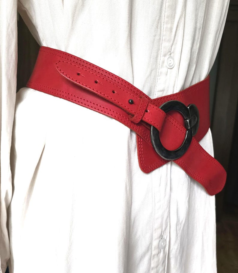 May include: A wide, red leather belt with a large, circular, silver-toned buckle. The belt is worn over a white shirt, showcasing its bold design and color. The belt's width is approximately 4 inches.