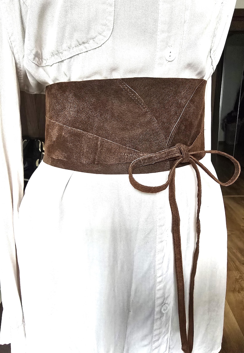 Puede incluir: Cintur&oacute;n ancho de ante marr&oacute;n con cierre de lazo. El cintur&oacute;n se lleva sobre una camisa blanca, creando un efecto de capas. La textura y el color del cintur&oacute;n contrastan con la camisa.