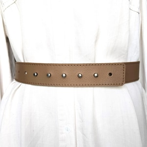 Op de afbeelding: Een beige leren riem met een reeks gelijkmatig verdeelde, zilverkleurige metalen studs langs de voorste band. De riem heeft een glad, gestructureerd oppervlak met zichtbare stiksels langs de boven- en onderranden.
