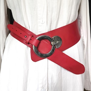 May include: A wide, red leather belt with a large, silver-toned buckle. The belt is designed to cinch the waist and features a unique, asymmetrical shape. The buckle is a prominent circular design.