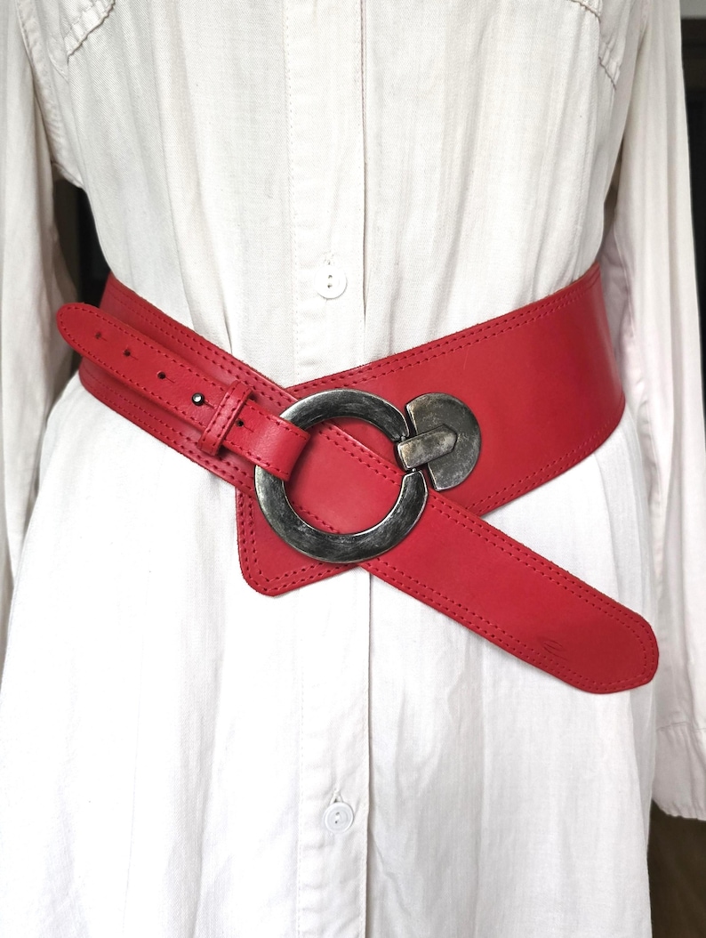 May include: A wide, red leather belt with a large, silver-toned buckle. The belt is designed to cinch the waist and features a unique, asymmetrical shape. The buckle is a prominent circular design.