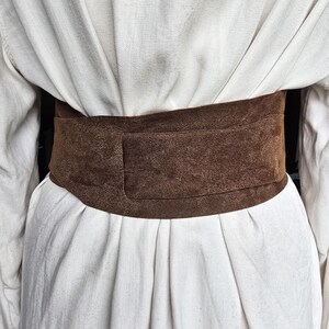 Puede incluir: Un cintur&oacute;n ancho de ante marr&oacute;n usado sobre un vestido blanco. El cintur&oacute;n es de color liso y parece estar hecho de un material suave y texturizado. El vestido tiene pliegues y un dise&ntilde;o sencillo.