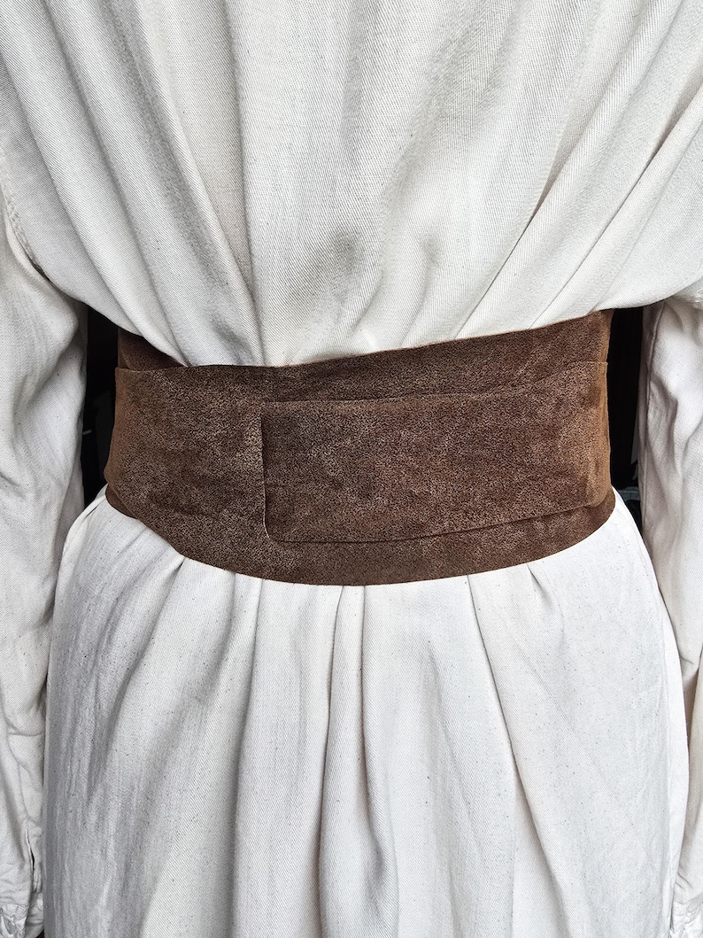 Puede incluir: Un cintur&oacute;n ancho de ante marr&oacute;n usado sobre un vestido blanco. El cintur&oacute;n es de color liso y parece estar hecho de un material suave y texturizado. El vestido tiene pliegues y un dise&ntilde;o sencillo.