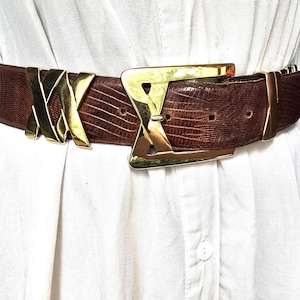 May include: A brown belt with a gold-coloured buckle and accents. The belt has a textured, reptile-like pattern. The buckle is a large, geometric design. The belt is worn over a white shirt.