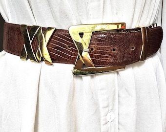 May include: A brown belt with a gold-colored buckle and accents. The belt has a textured, reptile-like pattern. The buckle is a large, geometric design. The belt is worn over a white shirt.