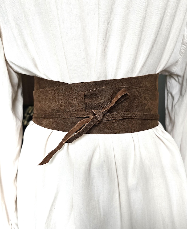 Puede incluir: Cintur&oacute;n ancho de ante marr&oacute;n atado en la parte delantera. El cintur&oacute;n est&aacute; envuelto alrededor de una prenda blanca. El cintur&oacute;n tiene un aspecto texturizado y una corbata larga y delgada. Accesorio de moda.
