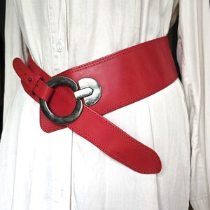 May include: A wide, red leather belt with a large, silver-toned buckle. The belt is cinched around a white shirt, showcasing its bold design and contrasting colors. The belt's width is approximately 4 inches.