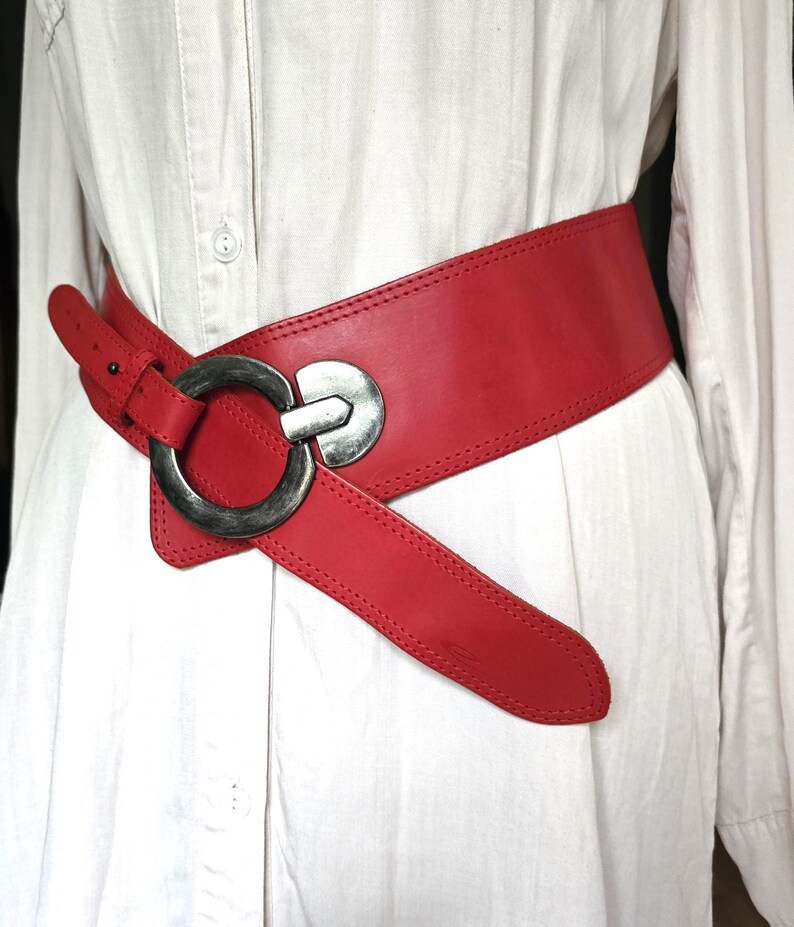 May include: A wide, red leather belt with a large, silver-toned buckle. The belt is cinched around a white shirt, showcasing its bold design and contrasting colors. The belt's width is approximately 4 inches.