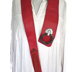 May include: A bold red leather belt with a large silver-tone metal ring buckle. The belt is worn over a white button-down shirt. The belt has a long, narrow strap with a buckle closure.