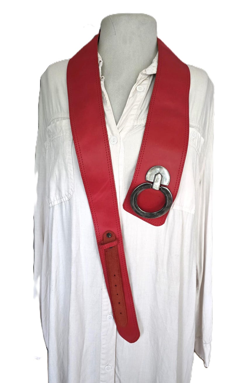 May include: A bold red leather belt with a large silver-tone metal ring buckle. The belt is worn over a white button-down shirt. The belt has a long, narrow strap with a buckle closure.