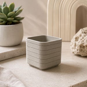 Può includere: Un vaso per piante di forma quadrata con una finitura grigio chiaro e dettagli a coste orizzontali lungo le pareti esterne. Il contenitore presenta una parte superiore liscia e aperta e una base piatta, con colore e consistenza uniformi.