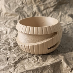 Può includere: Una ciotola sferica color beige stampata in 3D con un'ampia apertura superiore. L'esterno presenta una distinta texture a coste a spirale che avvolge la circonferenza, creando un aspetto stratificato con linee orizzontali visibili.