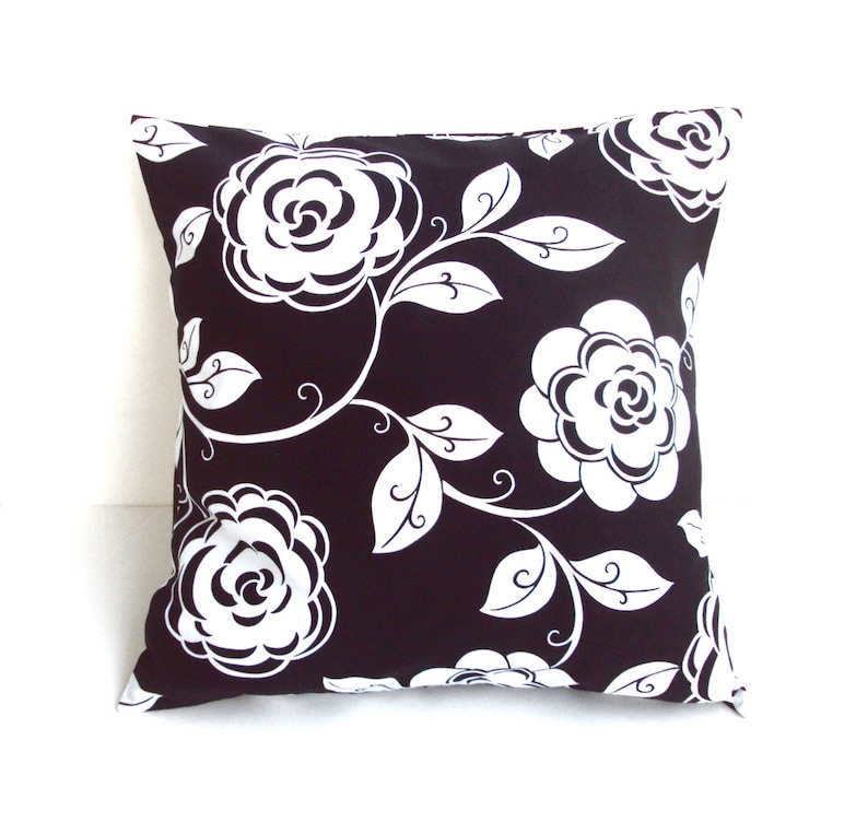 May include: Square decorative pillow with a black background and a white floral pattern. The design features large, stylized white roses and leaves with curved stems. The pillow is a square shape, perfect for a couch or bed.