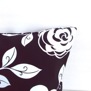 May include: Close-up of a decorative pillow with a black background and a white floral pattern. The design features large white roses and stylized leaves. The pillow's corner is visible against a white background.