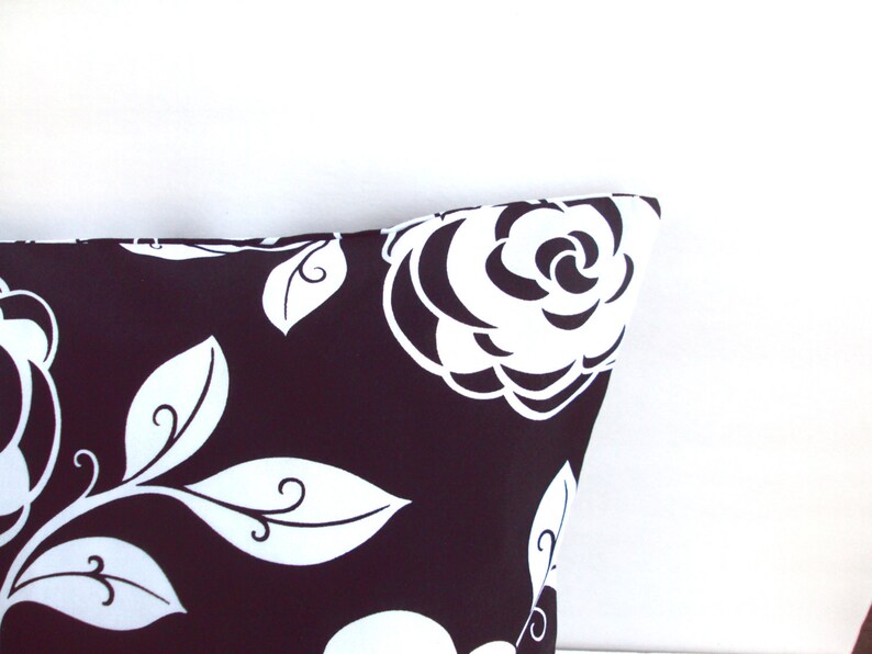May include: Close-up of a decorative pillow with a black background and a white floral pattern. The design features large white roses and stylized leaves. The pillow's corner is visible against a white background.