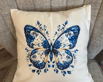 May include: A square off-white throw pillow featuring a large, centered butterfly graphic. The butterfly wings are decorated with intricate blue floral and vine patterns, with deep blue and light tan accents throughout the symmetrical design.