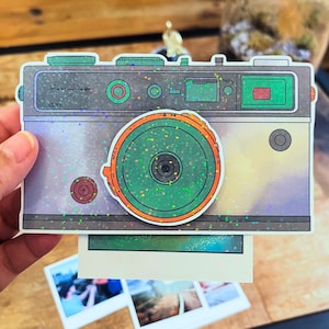 May include: A rectangular sticker featuring a vintage-style camera with a holographic finish. The camera body is grey with green accents, a prominent orange-rimmed circular lens, and small red details. A white photo print extends from the bottom.