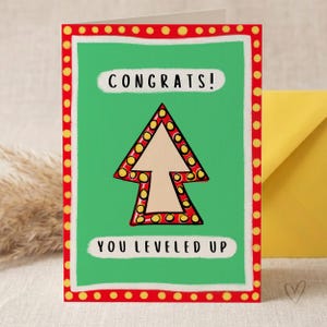 Peut inclure: Une carte de vœux avec un fond vert et une bordure rouge à pois jaunes. La carte présente une flèche beige pointant vers le haut, entourée de rouge avec des pois jaunes. Le texte indique CONGRATS! en haut et YOU LEVELED UP en bas.