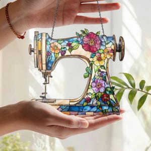 Könnte beinhalten: Buntglas-Nähmaschine mit floralen Akzenten. Das farbenfrohe Design zeigt eine blaue Basis, einen cremefarbenen Körper und eine Vielzahl von Blumen in Rosa, Lila, Gelb und Orange. Eine Kette ist zum Aufhängen befestigt.