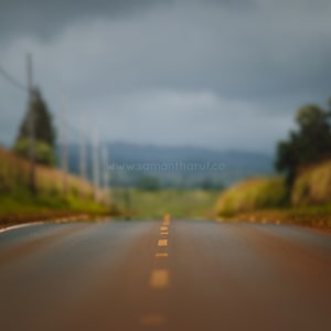 May include: A long, straight, paved road with yellow lines down the center, leading to a distant horizon. The road is surrounded by green grass and trees, and the sky is overcast with gray clouds.