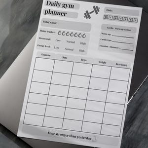Puede incluir: Un planificador diario de gimnasio blanco con un gráfico de mancuernas, sobre un portátil. El planificador incluye secciones para ejercicio, series, repeticiones, peso y descanso. También tiene un rastreador de agua, indicadores de nivel de estrés y energía.