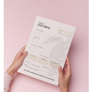 May include: A daily intentions planner with the text "TODAYS INTENTIONS" and a decorative turtle design. The planner includes sections for gratitude, goals, and a mood tracker. Held by hands against a pink background.