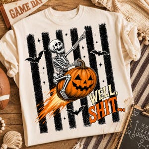 Puede incluir: Una camiseta de color crema con un gráfico de un esqueleto montado sobre una calabaza de Halloween como si fuera un cohete. El diseño incluye rayas verticales negras, murciélagos, estrellas y el texto naranja "WELL SHIT..." con una estela de fuego.