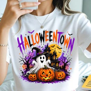 Puede incluir: Una camiseta blanca con un estampado gráfico que incluye el texto HALLOWEENTOWN en morado y naranja. El diseño muestra un fantasma blanco con sombrero de bruja negro, dos calabazas naranjas, una casa encantada oscura y una luna llena amarilla.