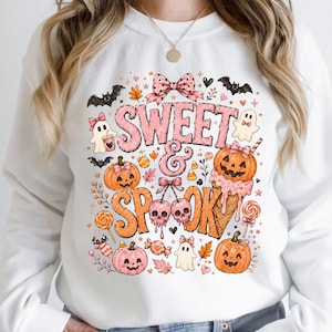 Puede incluir: Una sudadera blanca de manga larga con un estampado gráfico colorido que incluye el texto SWEET & SPOOK. El diseño presenta calabazas rosas y naranjas, fantasmas, murciélagos, dulces, cerezas y un cono de helado, decorados con lazos.
