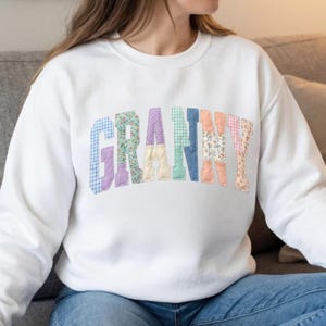 Puede incluir: Una sudadera blanca de cuello redondo con la palabra GRANNY en letras mayúsculas grandes y arqueadas. Cada letra está rellena con un patrón de tela de color pastel diferente, incluyendo texturas de cuadros, flores, lunares y lisas.