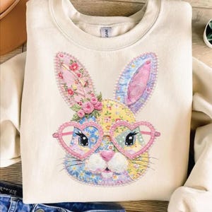 Puede incluir: Sudadera color crema con un rostro de conejo de retazos con gafas en forma de corazón. Las orejas y la cara del conejo están adornadas con estampados florales en rosa, azul y amarillo. Un racimo de rosas rosas se asienta sobre la cabeza del conejo.
