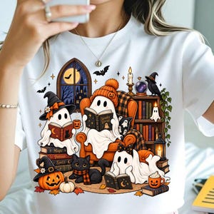 Puede incluir: Una camiseta blanca con un gráfico colorido de tres fantasmas leyendo libros en una habitación con estantería, una calabaza, un gato negro y una ventana que muestra una noche de luna. La ilustración incluye una vela y una bola de cristal.