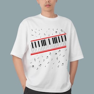 Puede incluir: Camiseta blanca con un diseño de teclado de piano. El teclado tiene teclas negras y blancas y líneas rojas. Notas musicales dispersas alrededor del gráfico. La camiseta tiene mangas cortas.