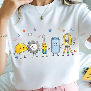 Puede incluir: Una camiseta blanca de manga corta con un gráfico colorido de cinco herramientas de construcción antropomórficas tomadas de la mano. Los personajes incluyen un casco amarillo, un engranaje gris, un destornillador, un plano azul y un teodolito amarillo.