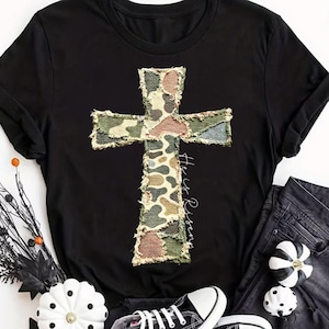Puede incluir: Camiseta negra con un diseño de cruz de camuflaje y la frase "He is Risen" en escritura cursiva. La cruz está hecha de parches de tela en tonos verdes, marrones y beige. La camiseta se combina con vaqueros negros y zapatillas.