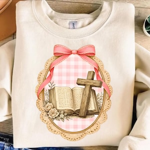 Puede incluir: Sudadera color crema con un lazo rosa y un gráfico enmarcado. El gráfico incluye una cruz de madera, un libro abierto y detalles florales sobre un fondo de cuadros rosas. El diseño está enmarcado con un borde festoneado.