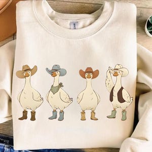 Może przedstawiać: Kremowa bluza z zabawną ilustracją czterech gęsi przebranych za kowbojów. Każda gęś ma na sobie kapelusz kowbojski i buty, a jedna również kamizelkę i apaszkę. Projekt jest umieszczony centralnie z przodu.