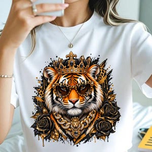 Puede incluir: Una camiseta blanca de manga corta con un estampado gráfico de un tigre que lleva una corona dorada, gafas de sol con estampado de leopardo y una gruesa cadena de oro. El tigre está rodeado de rosas negras y salpicaduras de pintura dorada.