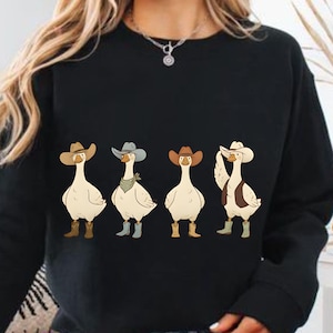 May include: Black sweatshirt with a cartoon design of four geese in cowboy attire. Each goose is depicted wearing a cowboy hat and boots, with one also sporting a vest and bandana. The design has a fun, country and western theme.